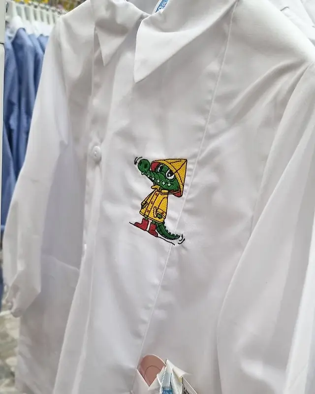 Grembiule bianco da bambino con colletto a punta e ricamo di un coccodrillo con impermeabile giallo